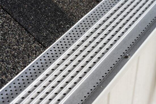 Close-up view of a Gutterglove stainless steel micro-mesh gutter guard with small round holes, installed along the edge of a roof with dark asphalt shingles. The device is designed to keep debris out of the gutter.