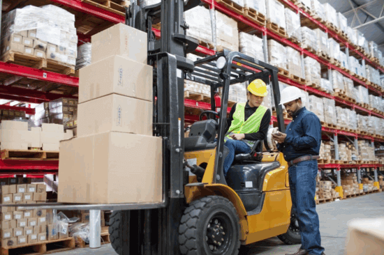 A worker uses a forklift to move stacked boxes in a warehouse featuring RiserStep Technology, while another worker stands nearby showing something on a clipboard. Both wear safety helmets and high-visibility vests.