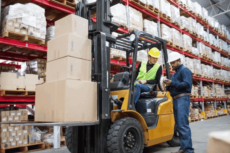 A worker uses a forklift to move stacked boxes in a warehouse featuring RiserStep Technology, while another worker stands nearby showing something on a clipboard. Both wear safety helmets and high-visibility vests.