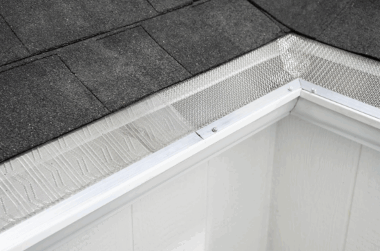 Close-up of a roof corner with dark shingles and a gutter guard featuring V-Bend Technology, installed over a white rain gutter to prevent debris from entering. The siding below the gutter is white.